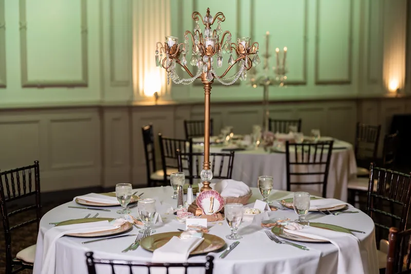 A round banquet table dressed in white linen holds a tall candelabra with pink flowers, place settings, and glassware beneath