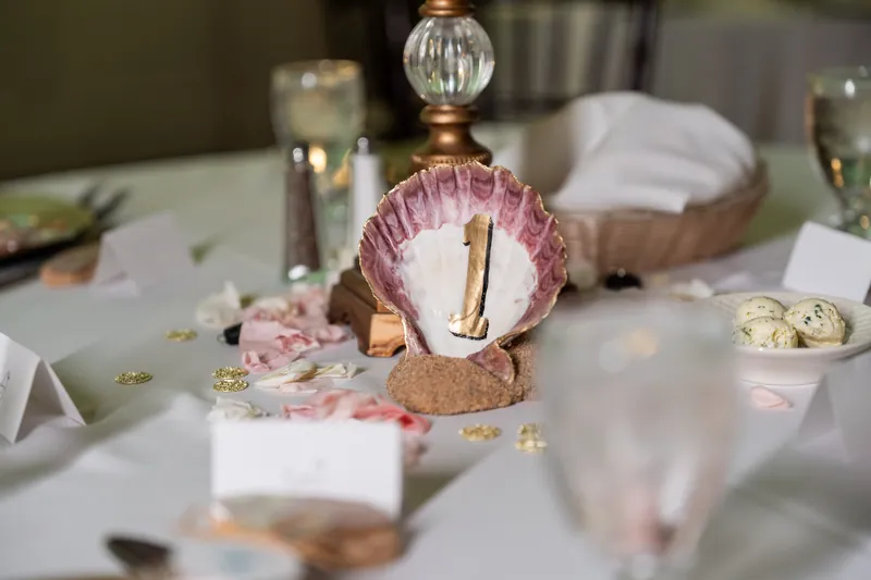 A table number sits in a pink shell-shaped holder surrounded by scattered gold confetti and glassware on a white tablecloth.