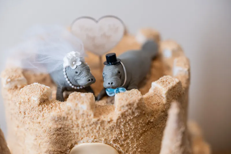 Two decorative shoes topped with small heart ornaments sit nestled in sand on a cake featuring a beach and ocean theme.