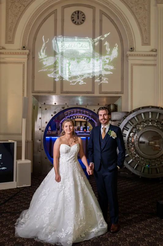 Sybil in a beaded white gown and Chris in a dark suit stand beneath an arched doorway with blue neon signage at The Treasury