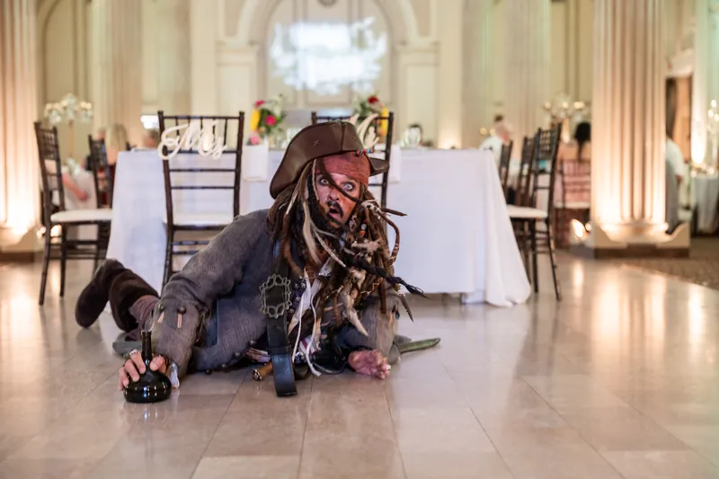 A costumed performer crawls across the polished ballroom floor in character, with white-draped tables and classical
