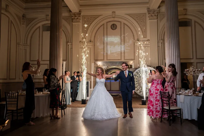 Sybil and Chris dance together under a glowing floral arch while family and friends look on in the grand columned ballroom.