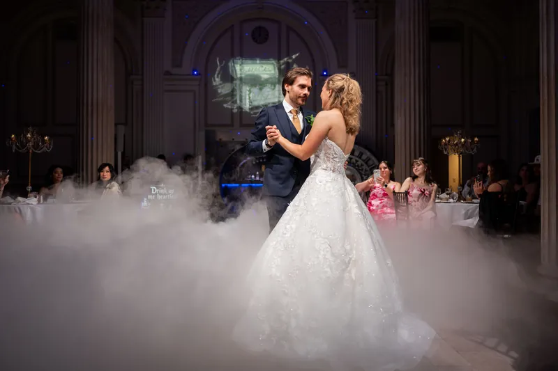 The couple sways together shrouded in fog during their first dance, with attendants and architectural columns framing