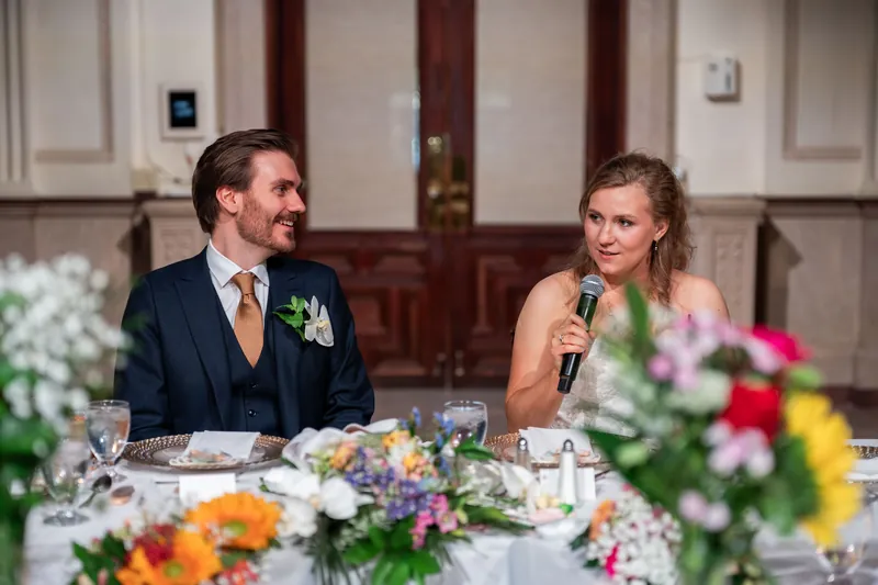 Chris speaks at the reception table while Sybil listens beside him, with colorful floral arrangements and place settings