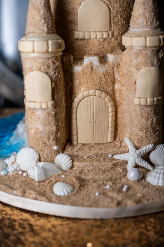 Tiered sand castle cake features detailed towers, arched doorways, and white seashell embellishments on its base.