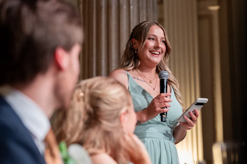 A woman in a sage-green dress speaks into a microphone while addressing guests during the reception.