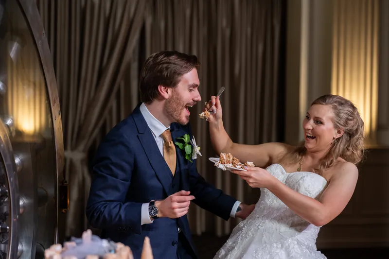 Sybil in a white strapless gown feeds Chris a bite of cake while he laughs during their reception.