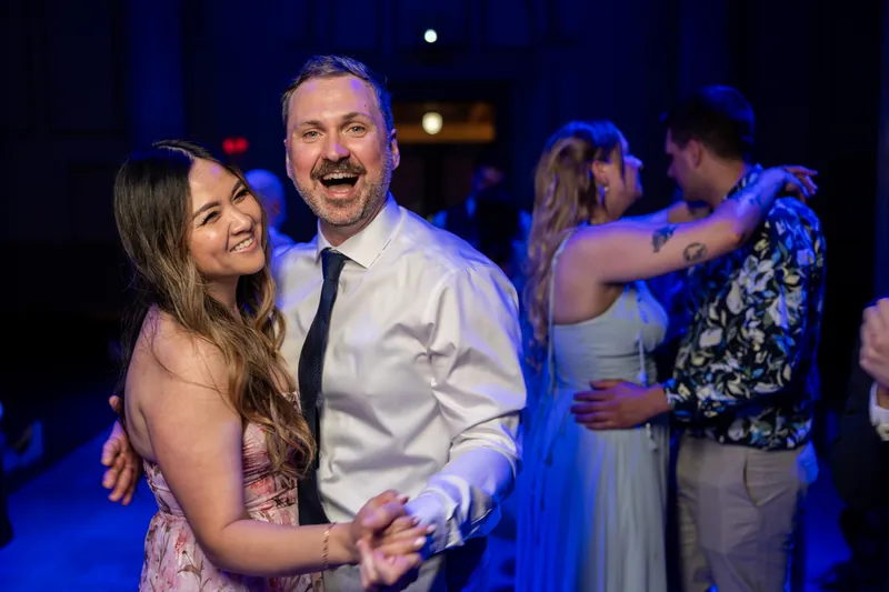 Sybil and Chris laugh while dancing, with other guests visible dancing behind them in the blue-lit ballroom.