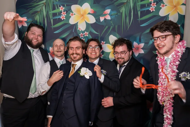 Chris and five groomsmen stand in front of a tropical floral backdrop, celebrating with gestures and lei-style garlands.