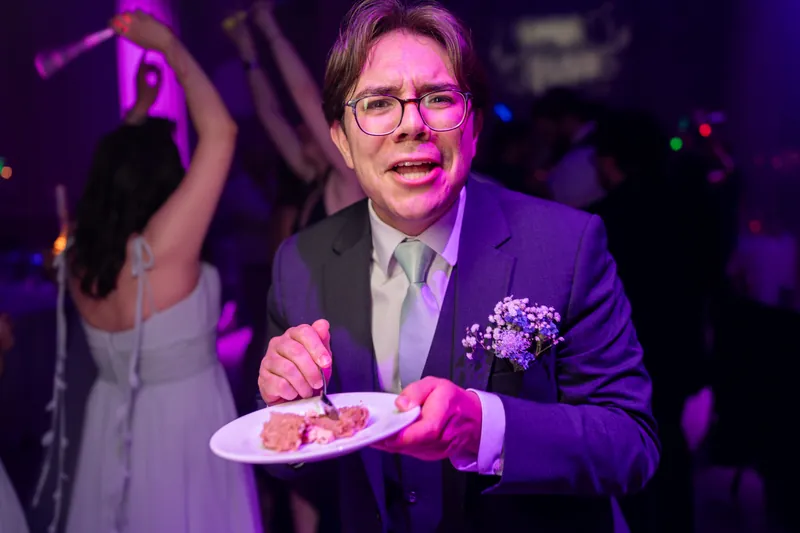Chris in a purple blazer holds a plate of food under vibrant pink lighting during the reception.