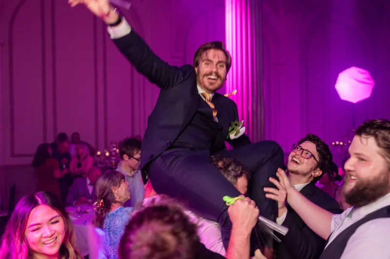 Chris is lifted onto guests' shoulders with his arm raised, celebrating with the crowd in purple-lit surroundings.