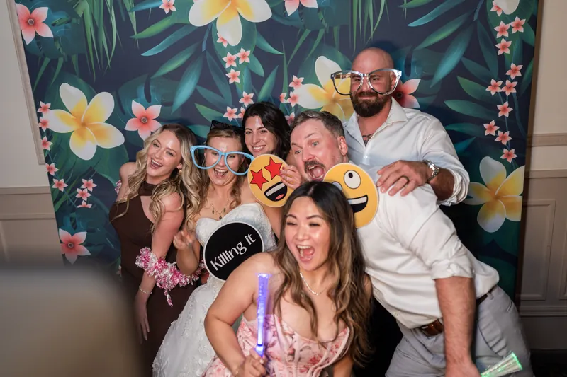 A group of wedding guests poses playfully in front of a tropical floral backdrop holding oversized photo booth props.