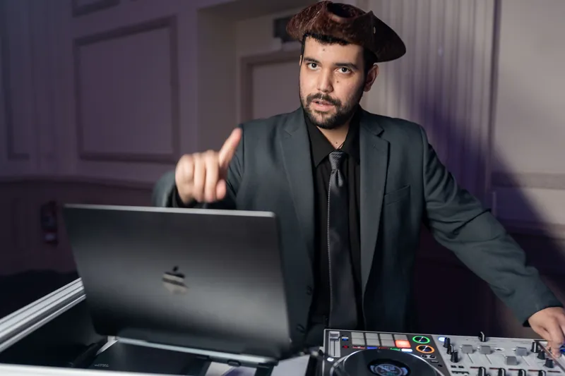 A DJ in a dark suit and beret operates equipment, adjusting settings on a laptop during the event.