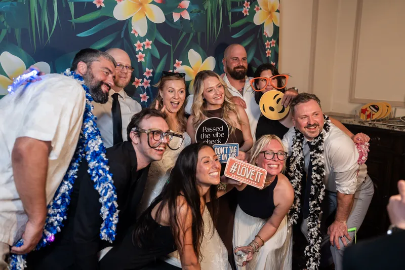 Guests cluster together in front of the floral photo booth backdrop, holding prop signs and grinning at the camera.