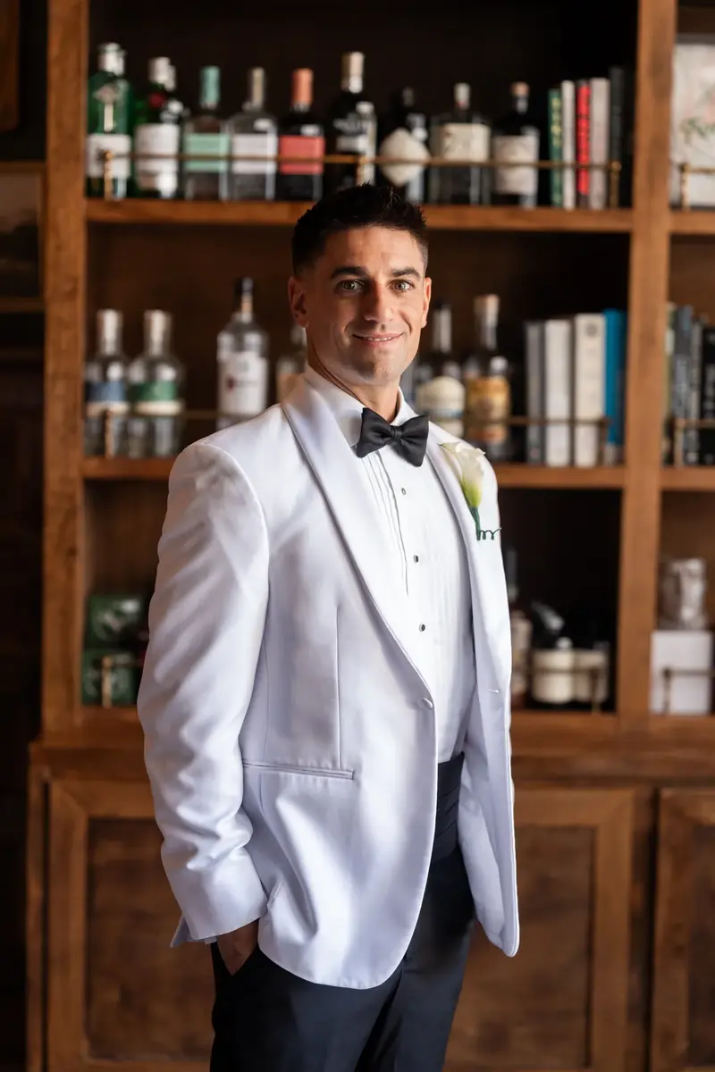 Jay portrait in white dinner jacket standing in front of bar shelves