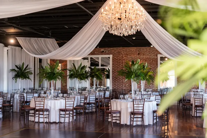 Reception room with crystal chandelier, draping, exposed brick, and palm arrangements