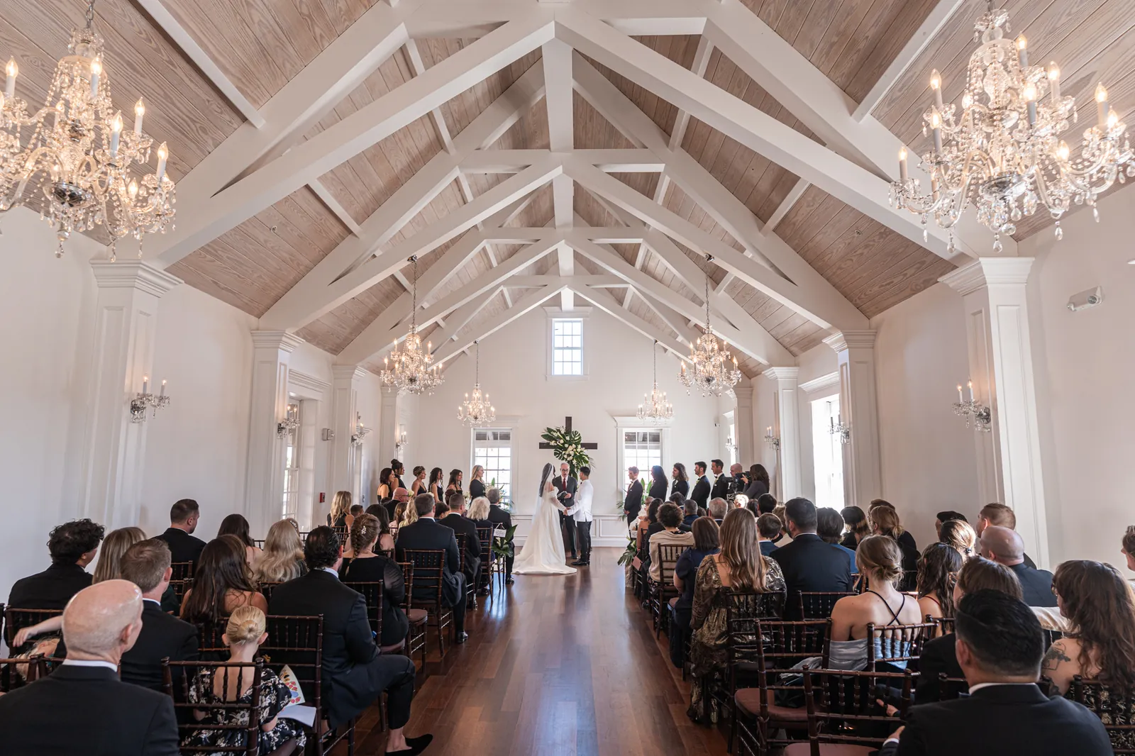 Rows of guests seated on either side of the aisle face a white vaulted ceiling with chandeliers during the ceremony.