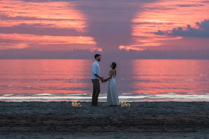 Thomas and Colleen hold hands on Vilano Beach at sunset, candles flickering in the wet sand below a pink and purple sky.