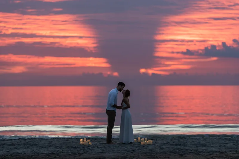 Thomas kneels before Colleen on the sand at sunset while candles flicker around them and vibrant pink clouds fill the sky.