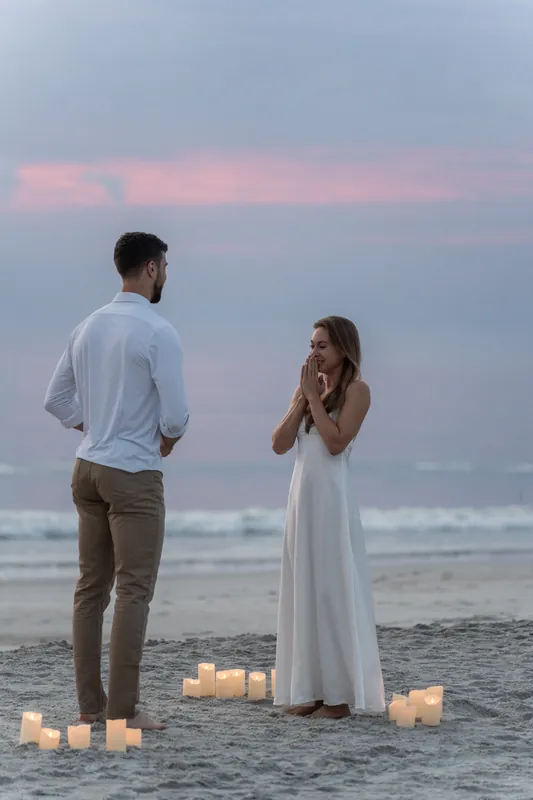 Colleen's hand covers her mouth in surprise as Thomas kneels on the beach, candlelight glowing around them at dusk.