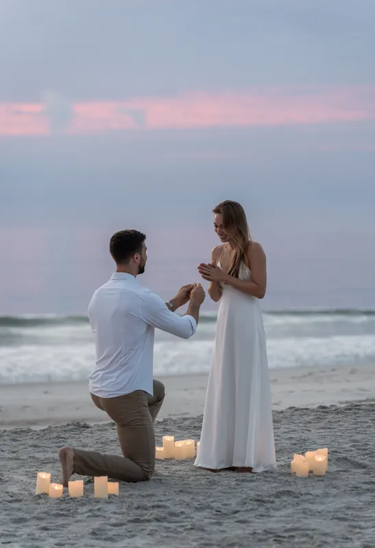 Colleen looks down at her ring as Thomas kneels before her on the sand, surrounded by glowing candles and soft twilight.