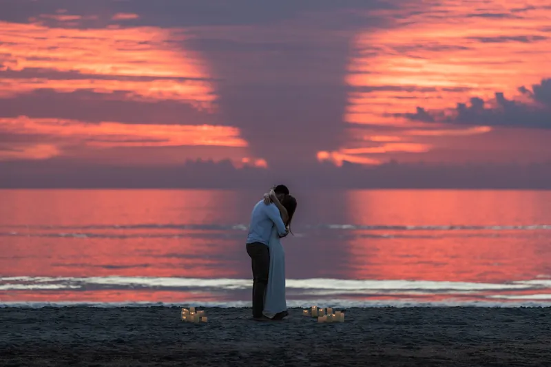 Thomas and Colleen embrace at sunset on a beach as dramatic orange and pink clouds fill the sky above the ocean.