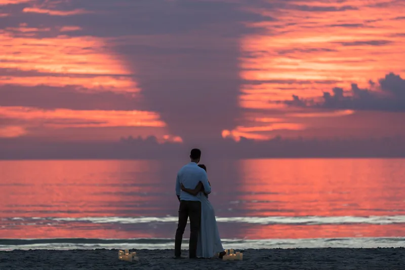 Colleen stands alone facing the water at dusk, wearing a white dress while vibrant clouds reflect across the horizon.