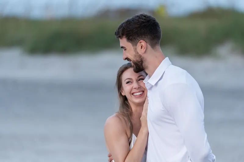 Thomas and Colleen smile together near the shoreline, with marsh grasses visible behind them under soft evening light.
