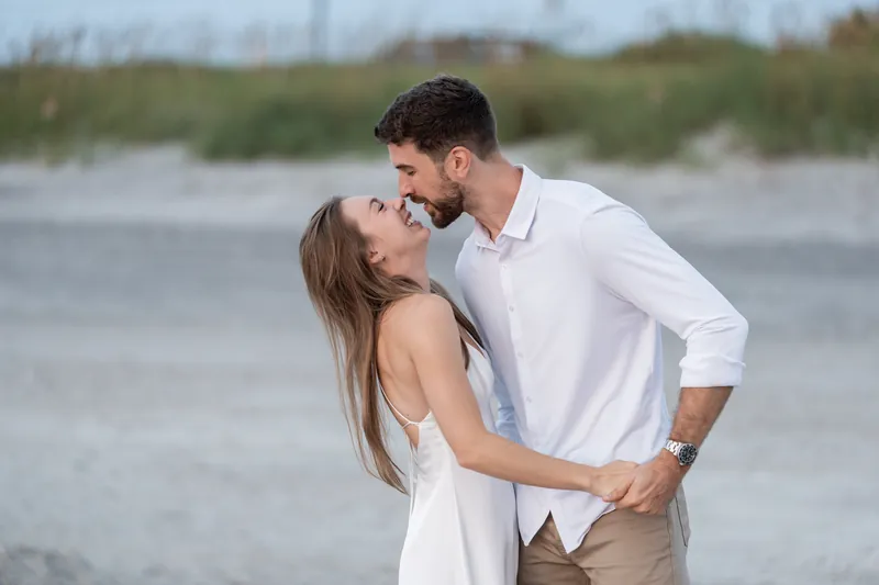 Thomas and Colleen kiss on the sand at Vilano Beach with ocean and dunes behind them.
