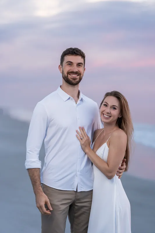 Thomas and Colleen stand together at dusk as Colleen displays her engagement ring with a smile.