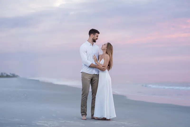 Thomas holds Colleen close on the sand during golden hour, the couple framed against a pastel sky and calm ocean behind them.