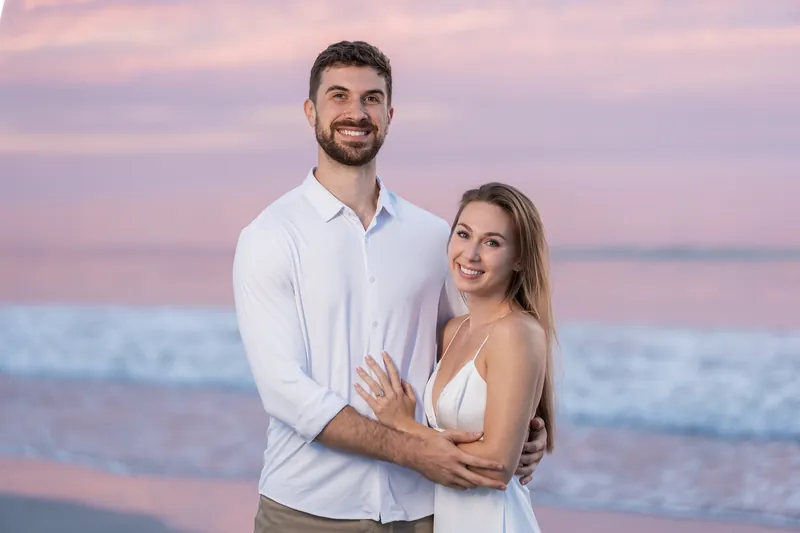 Thomas and Colleen embrace at sunset on Vilano Beach with pink and purple skies stretching across the horizon behind them.