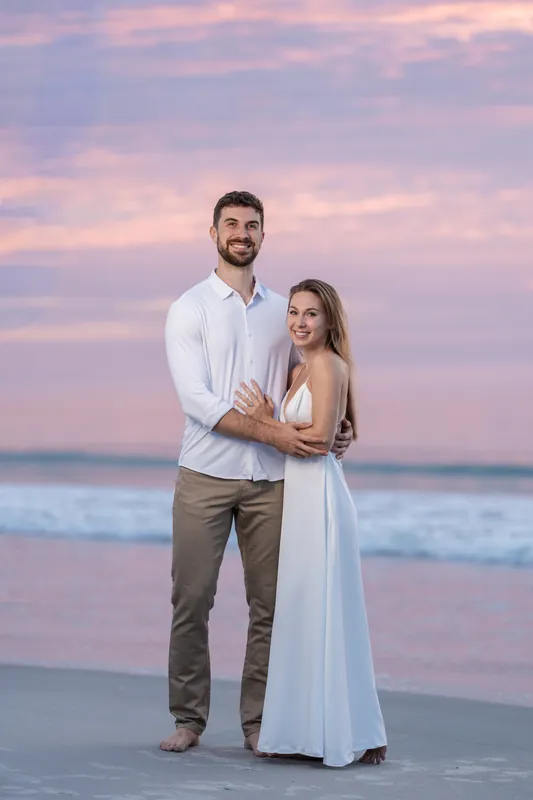 Thomas and Colleen embrace in the sand at Vilano Beach as waves lap nearby under a purple-pink sunset sky.