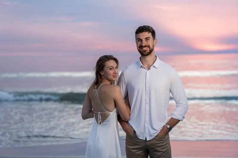 Thomas rests his hand on Colleen's back as they stand together on the beach with golden light overhead.