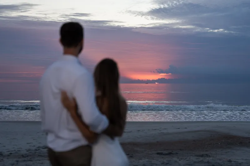 Thomas and Colleen embrace softly while an orange and pink sunset glows across the water behind them.