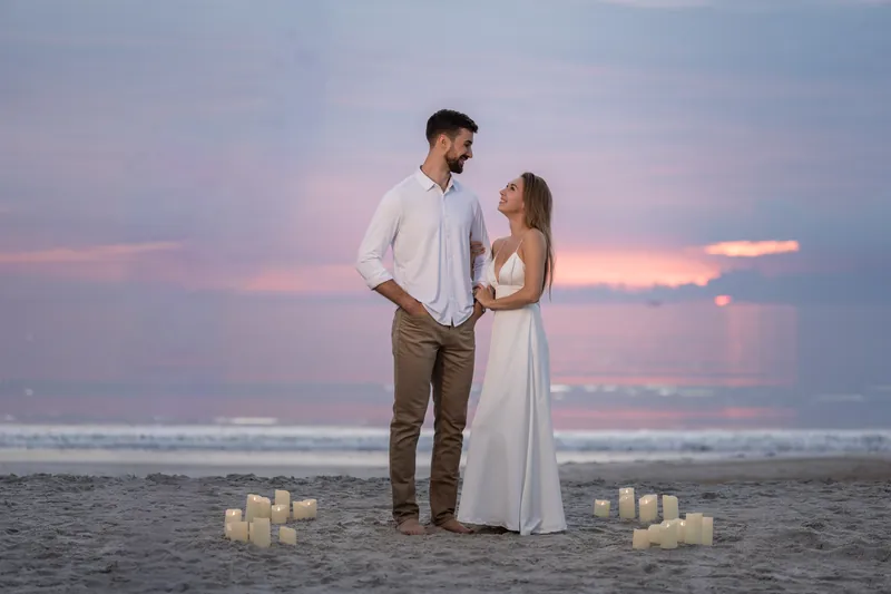 Thomas and Colleen stand surrounded by lit candles on the sand as the sunset casts warm light across the sky.