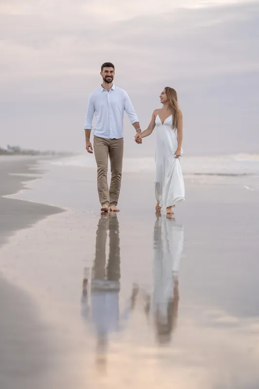Thomas and Colleen walk hand in hand across wet sand at Vilano Beach, their reflections mirrored below.