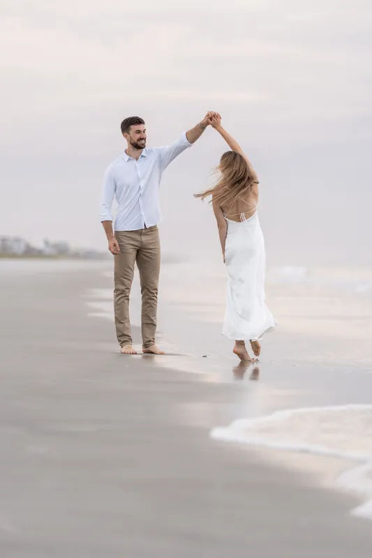 Colleen twirls with her arm raised as Thomas watches, both standing in the surf with the ocean behind them.