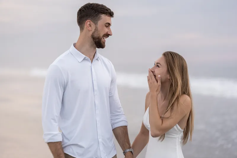 Smiling at each other, Thomas and Colleen stand close together on the beach with their hands linked.