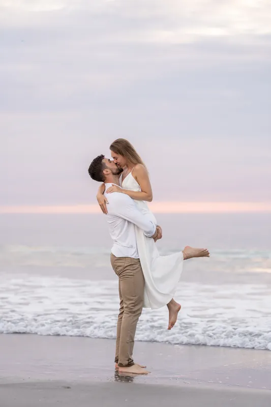 Thomas lifts Colleen into his arms while standing in the shallow surf, both laughing as the sun sets over the water.