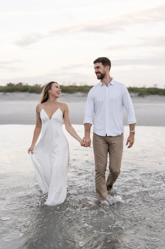 Thomas and Colleen hold hands wading through shallow water at Vilano Beach with an overcast sky above.