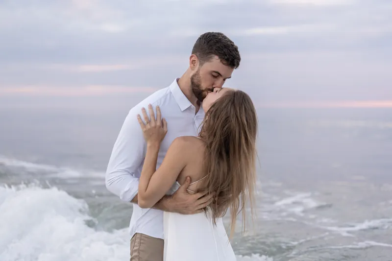 Thomas embraces Colleen from behind as ocean waves crash nearby, both facing the water at sunset.