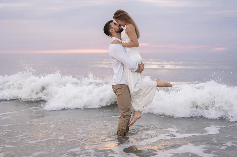 Thomas lifts Colleen in his arms while standing in the surf, the two kissing with waves breaking around them.