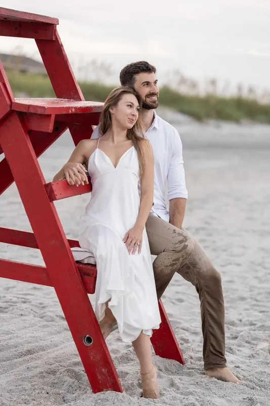 Colleen and Thomas lean against a large red lifeguard chair on the sand, looking out toward the water together.
