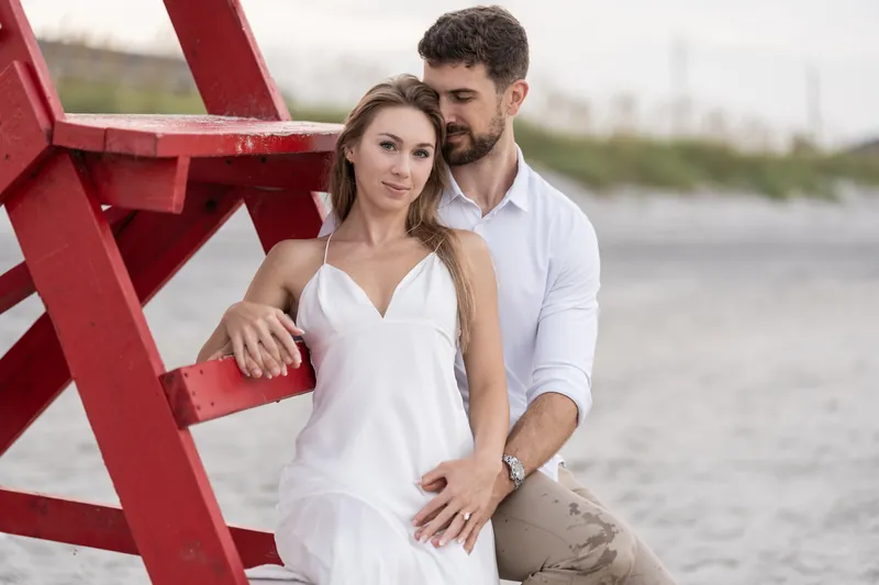 Thomas stands beside Colleen at the red lifeguard stand, his arm around her shoulders as they gaze toward the horizon.