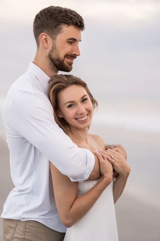 Thomas wraps his arms around Colleen from behind on the beach, both smiling softly toward the camera.