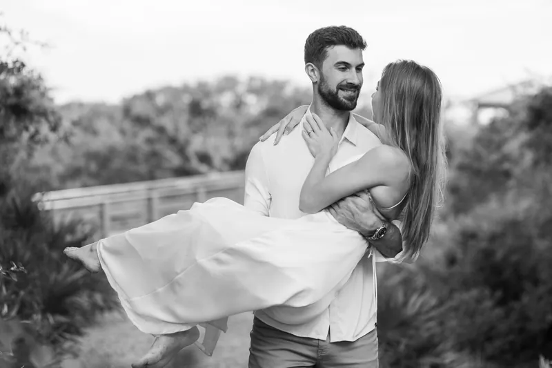 Thomas lifts Colleen in a black-and-white moment, both laughing with coastal landscape behind them.
