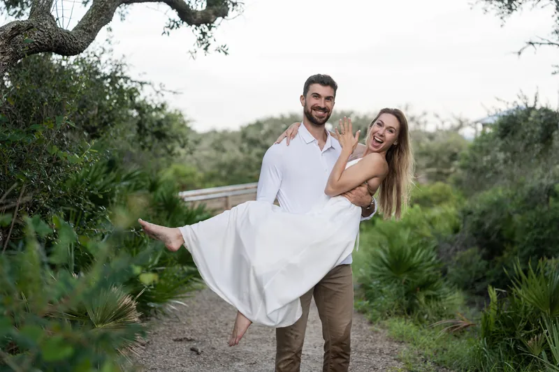 Thomas carries Colleen's flowing white dress as they pose on a pathway surrounded by lush greenery.