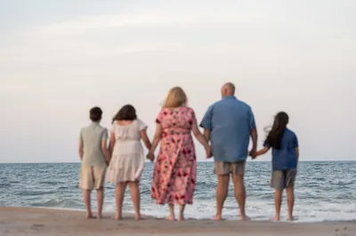 Trena's family of five stands in the shallow surf at St. Augustine Beach, holding hands and looking toward the water.