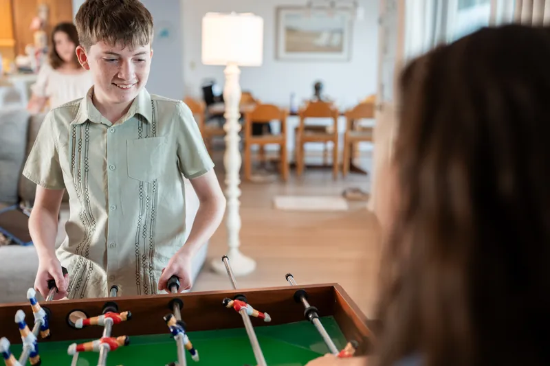 Boy in sage green button-up shirt smiles while playing foosball indoors.